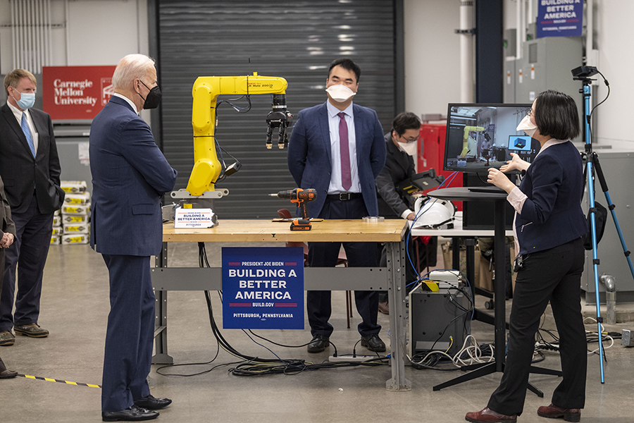  Joe Biden, in a blue suit, is shown standing to the left of a table, behind which a dark-haired man wearing a blue shirt, blue pants and mauve tie stands. A woman with dark hair is on the right, and a robotic arm is in the background.