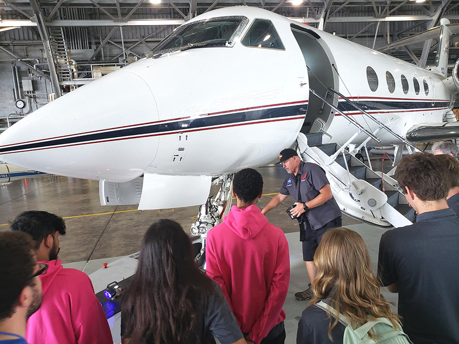  Students with their backs to the camera look at a small airplane in a hanger. A man in dark shorts, a button-down short-sleeved shirt and a hat points to an area near the plane's wheel.