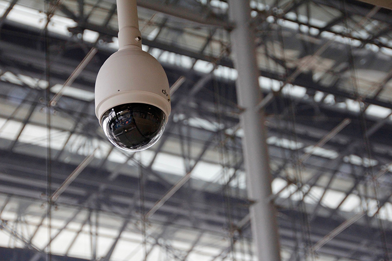  A video camera hangs from a white post on an industrial ceiling.