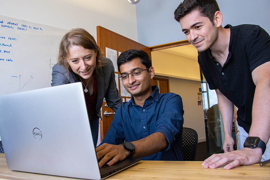  A man in the center of the photo works on a laptop while a woman on the left and man on the right look over his shoulder.