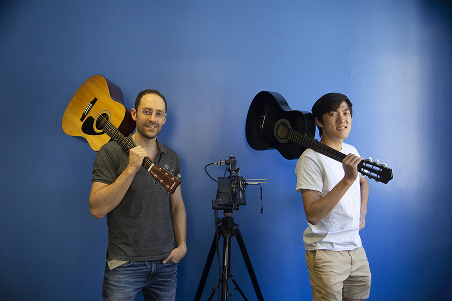  Two men separated by equipment on a tripod pose with guitars resting on their shoulders.