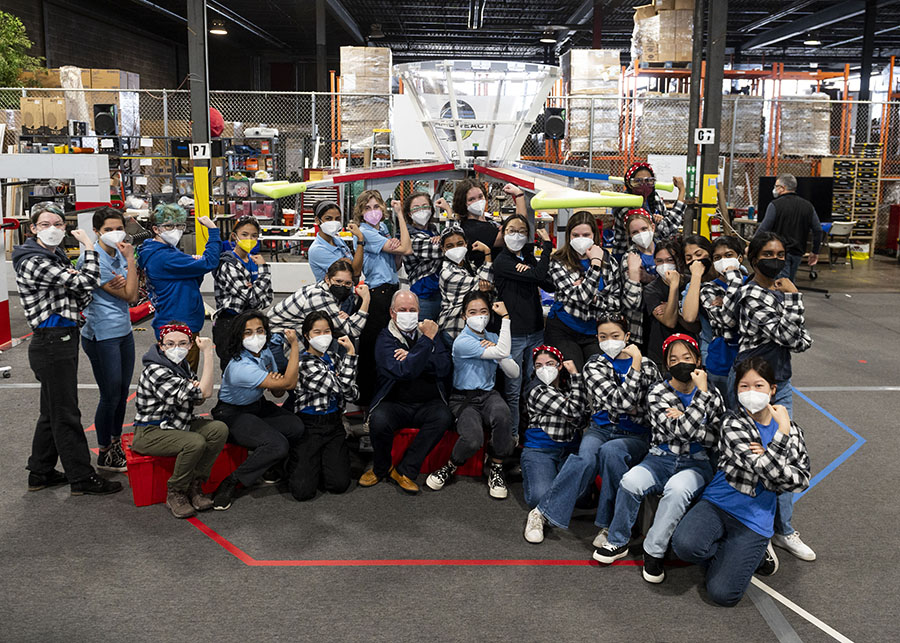  A group of young women in black and white plaid hoodies (some wearing bandanas in their hair) pose in the Rosie the Riveter stance with Congressman Mike Doyle against an industrial background.