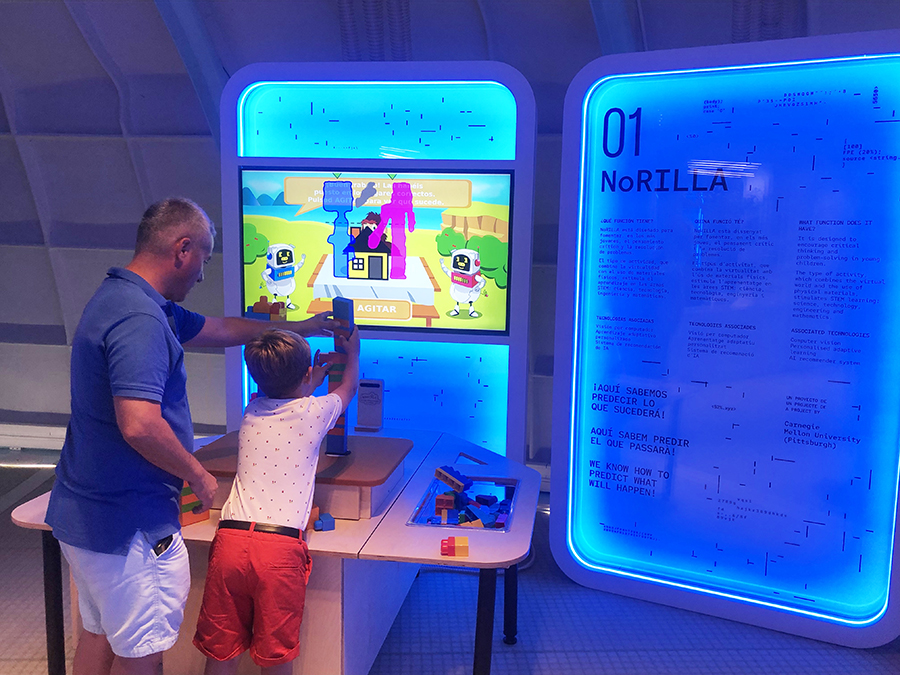  A man and child build a tower from blocks on a table, while a screen in the background shows a digital version of the tower and a gorilla that gives advice for making the tower stable.