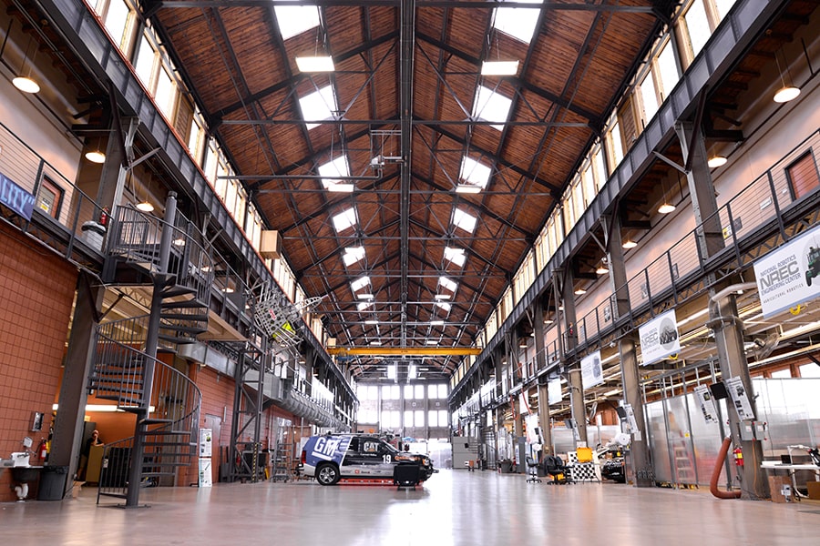  The Highbay at NREC: a long industrial room with a vaulted ceiling. A spiral staircase in the foreground connects the main floor to the second floor. An autonomous SUV is parked in the middle of the main floor.