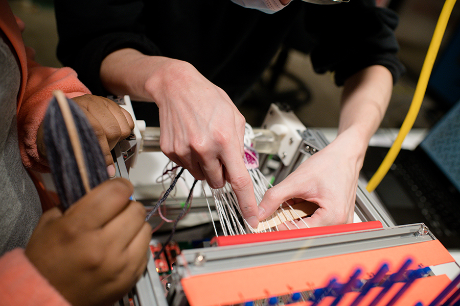  Two sets of human hands interact with fabric on a small robotic loom.