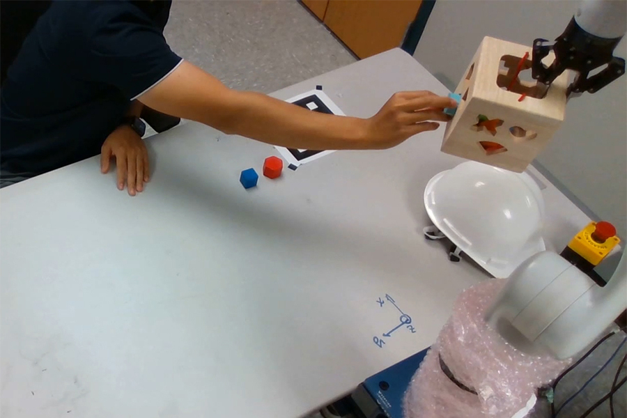  A human arm reaches across a table to insert a square-shaped block into the corresponding hole in a box held by a robotic hand.