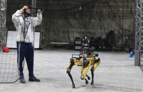  A man on the left of the photo interacts with a four-legged robot in an underground environment. The drone is yellow with black spots.