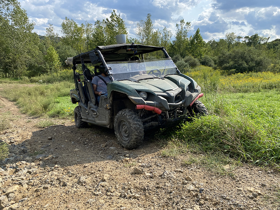  A green all-terrain vehicle outfitted with sensors sits half in the grass and half on a dirt road. Two people sit inside the ATV.